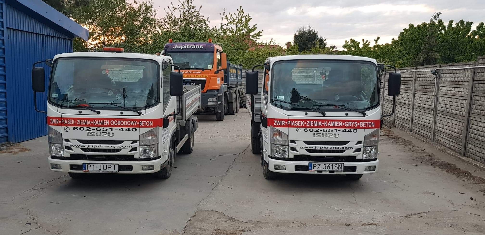 Flota białych samochodów ciężarowych firmy transportowej z pomarańczowym samochodem ciężarowym w tle, oferującej transport materiałów budowlanych, z widocznym numerem telefonu i adresem strony...