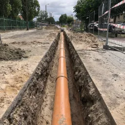 Pomarańczowa rura kanalizacyjna ułożona w wykopie wzdłuż ulicy, zabezpieczona barierkami, z widocznymi narzędziami i stertami ziemi po bokach.