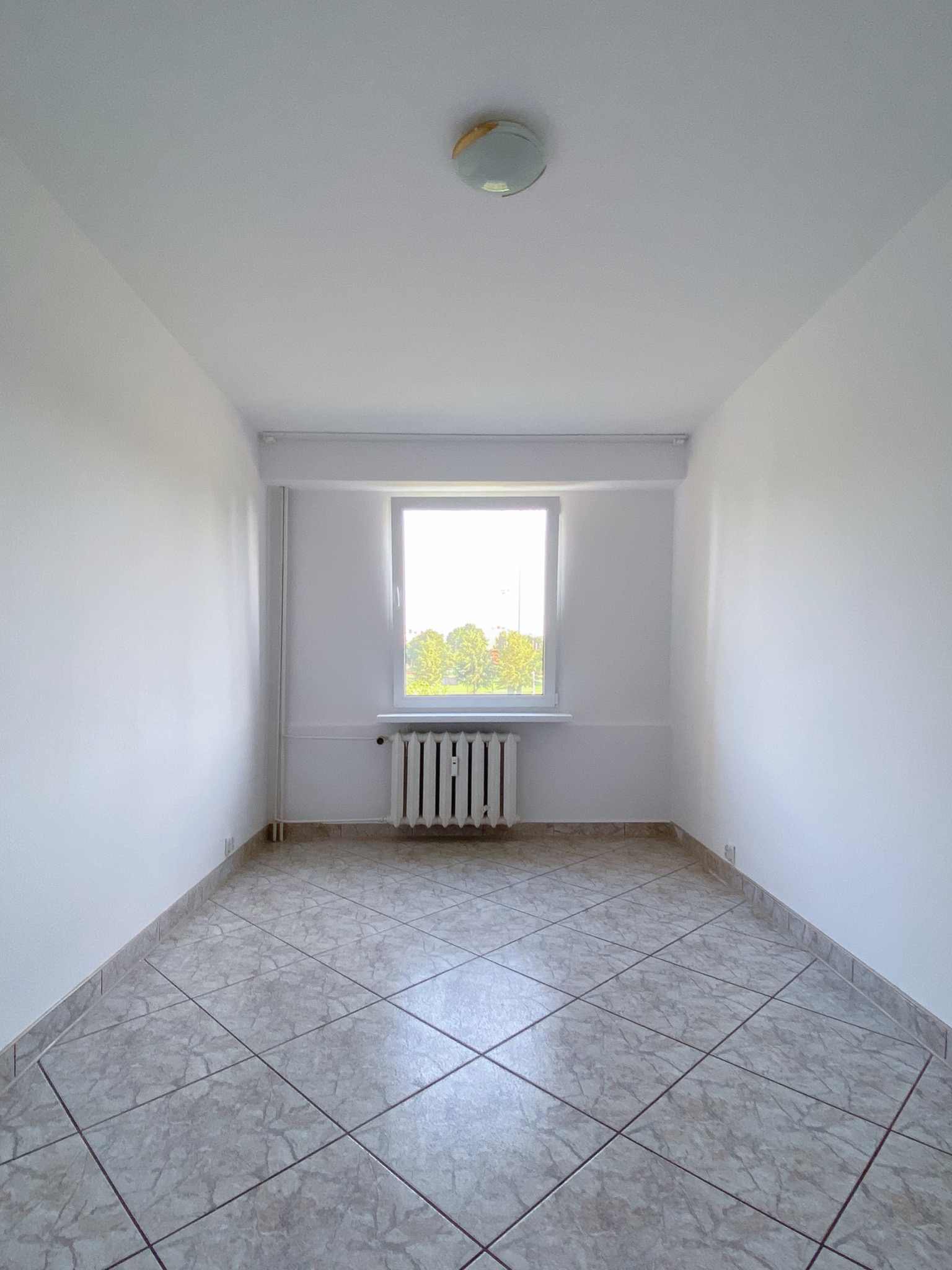 Puste, wyremontowane pomieszczenie z białymi ścianami, grzejnikiem pod oknem i podłogą wyłożoną płytkami w odcieniach szarości i beżu.