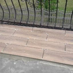 Układanie płytek imitujących drewno na balkonie, widoczne krzyżyki dystansowe i zaprawa, w tle ozdobna balustrada i zieleń.