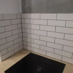 Białe, prostokątne płytki ułożone na ścianie nad blatem kuchennym z czarną płytą indukcyjną, szare fugi, surowy beton na g&oacute;rze ściany.