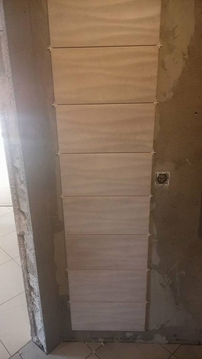 Układanie podłużnych, beżowych płytek ceramicznych na ścianie, z użyciem krzyżyków dystansowych, widoczny surowy stan ściany i podłogi.