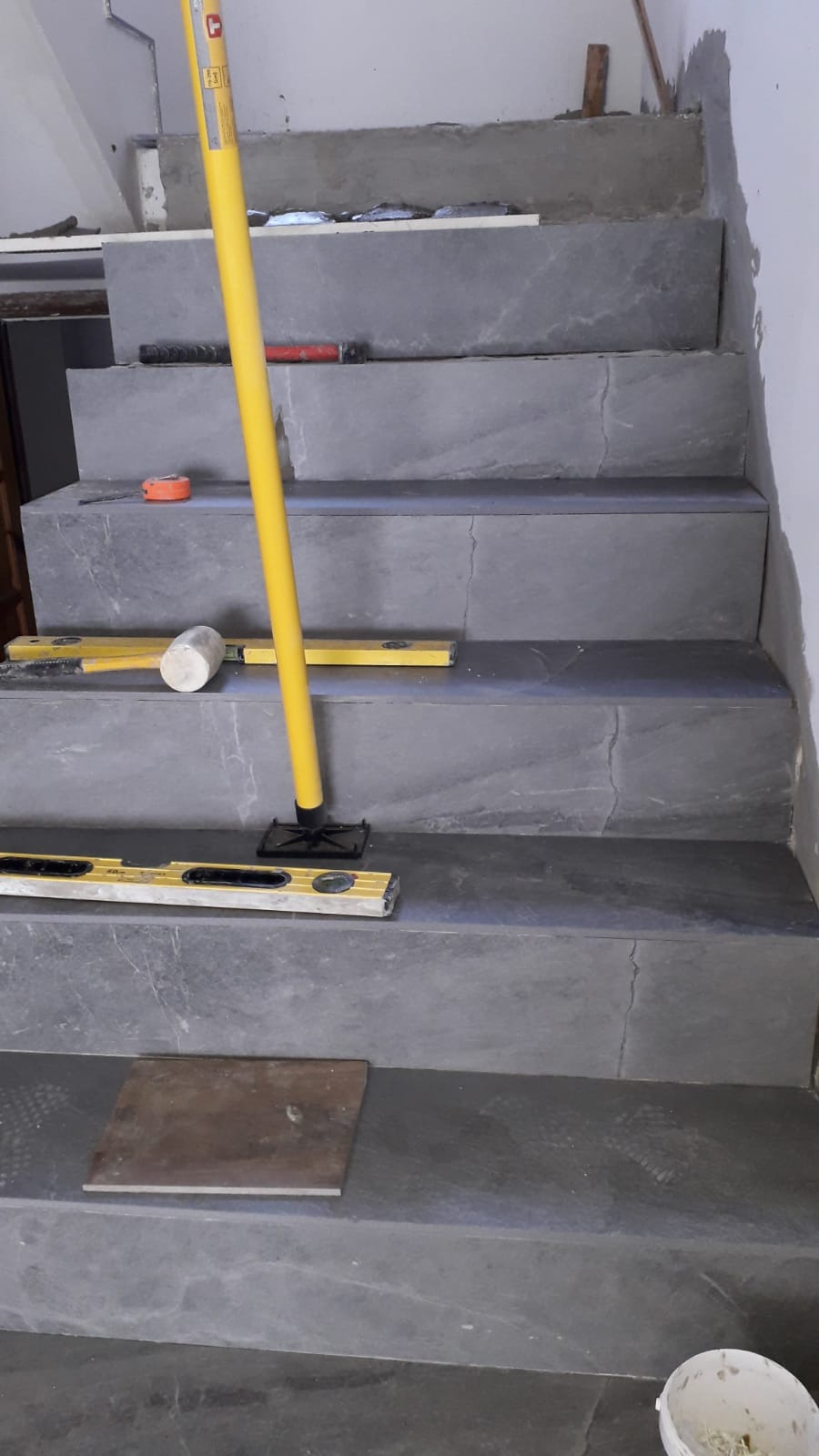 Schody wyłożone szarymi płytkami imitującymi kamień, w trakcie montażu widoczne poziomice, gumowy młotek i inne narzędzia glazurnicze.