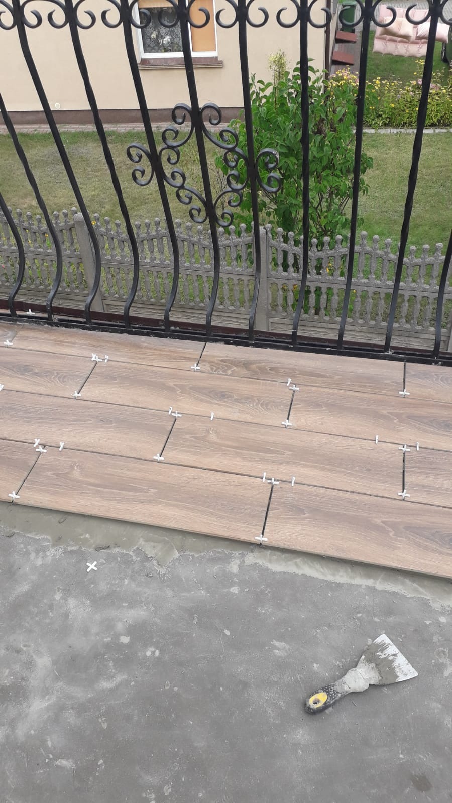 Układanie płytek imitujących drewno na balkonie, widoczne krzyżyki dystansowe i zaprawa, w tle ozdobna balustrada i zieleń.