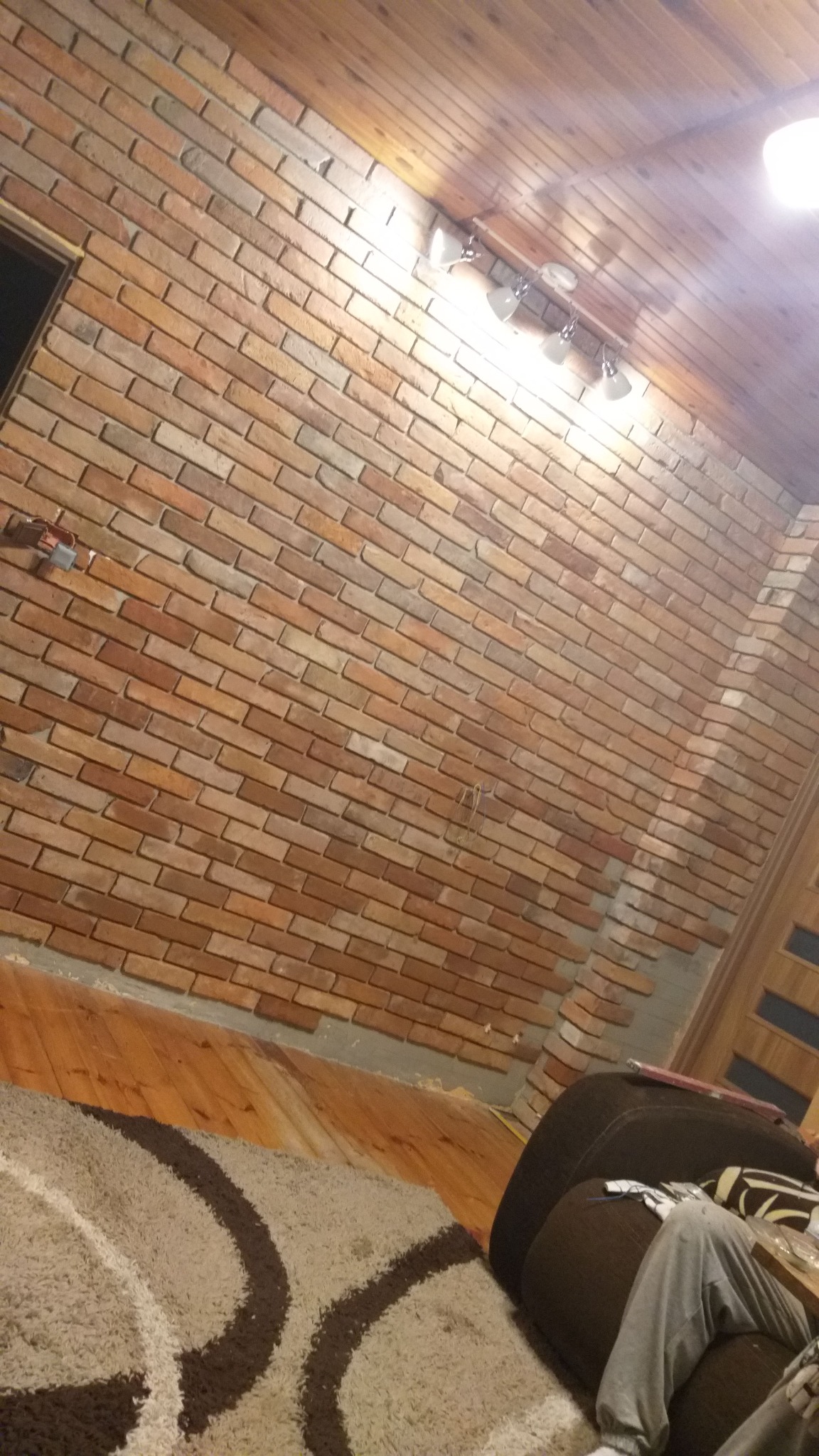 Ceglana ściana w trakcie remontu, widoczne niedokończone fragmenty przy podłodze, drewniany sufit z oświetleniem punktowym, dywan z geometrycznym wzorem na podłodze.