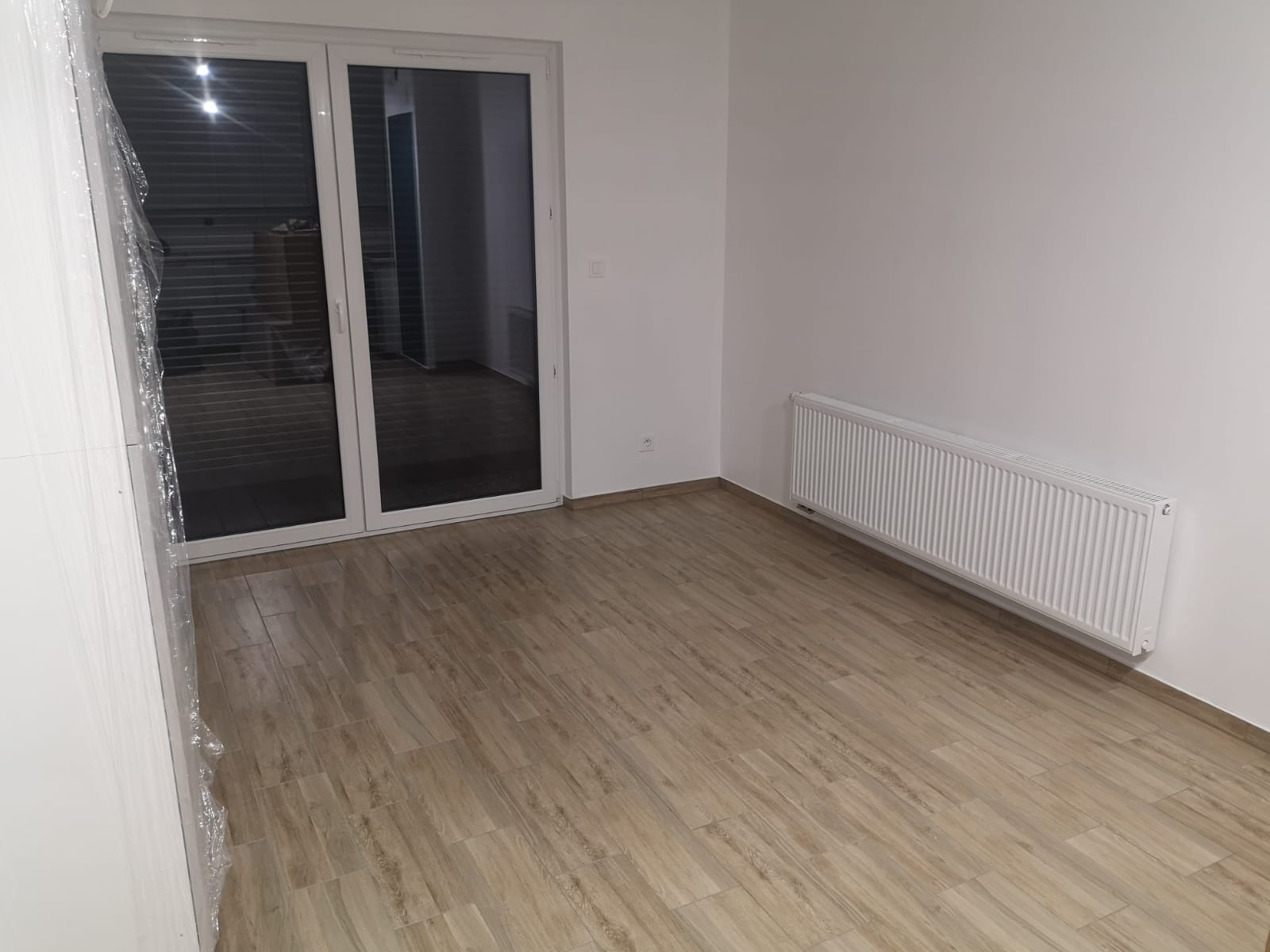 Puste pomieszczenie z panelami podłogowymi imitującymi drewno, białym grzejnikiem i oknem balkonowym z białymi ramami, częściowo owiniętym folią ochronną.