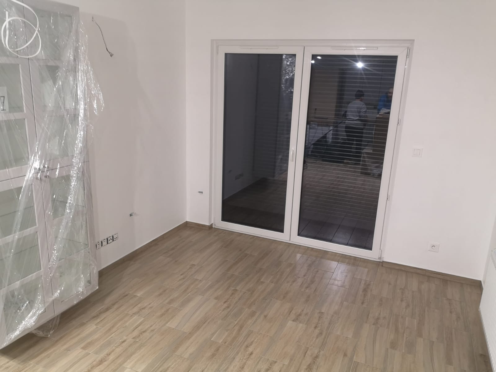 Pusty pokój z nową podłogą imitującą drewno, białe ściany i przesuwne drzwi balkonowe z żaluzjami; szafka okryta folią ochronną.