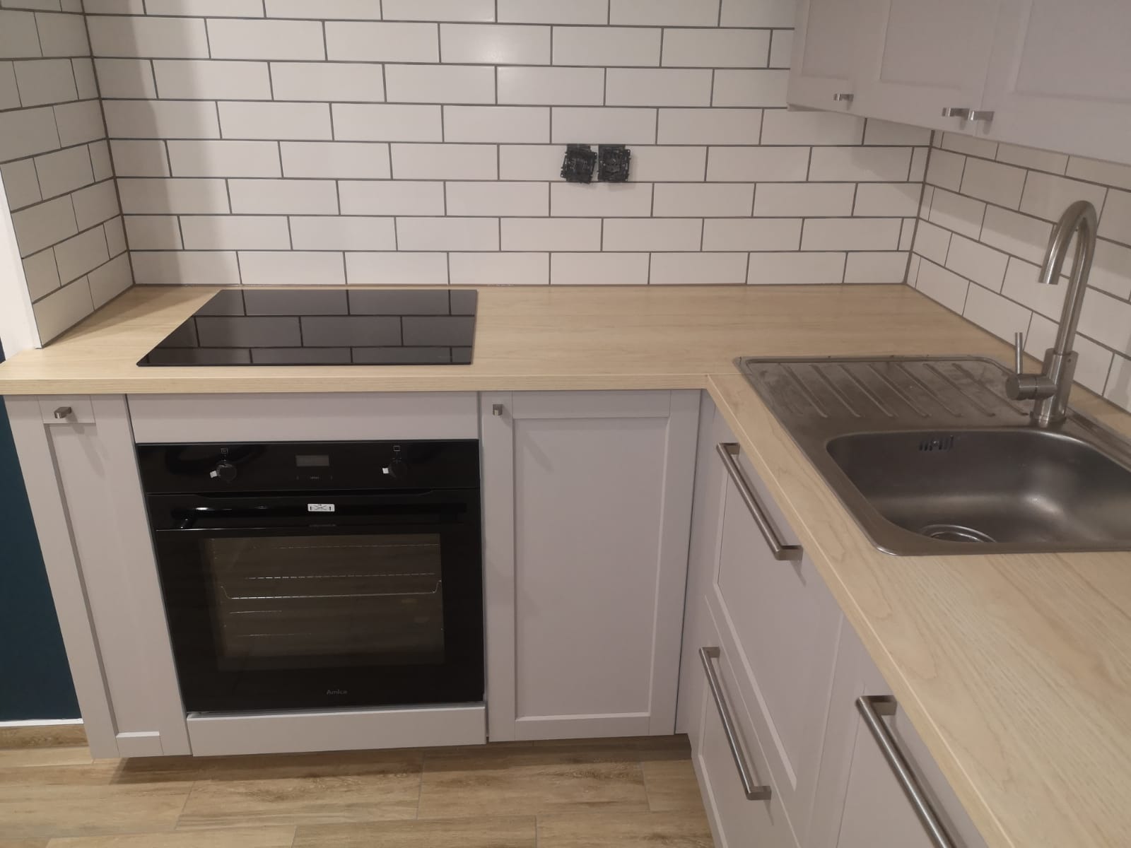 Nowoczesna kuchnia z białymi kafelkami ułożonymi w cegiełkę, jasnym drewnianym blatem, szarymi szafkami i wbudowanym piekarnikiem.