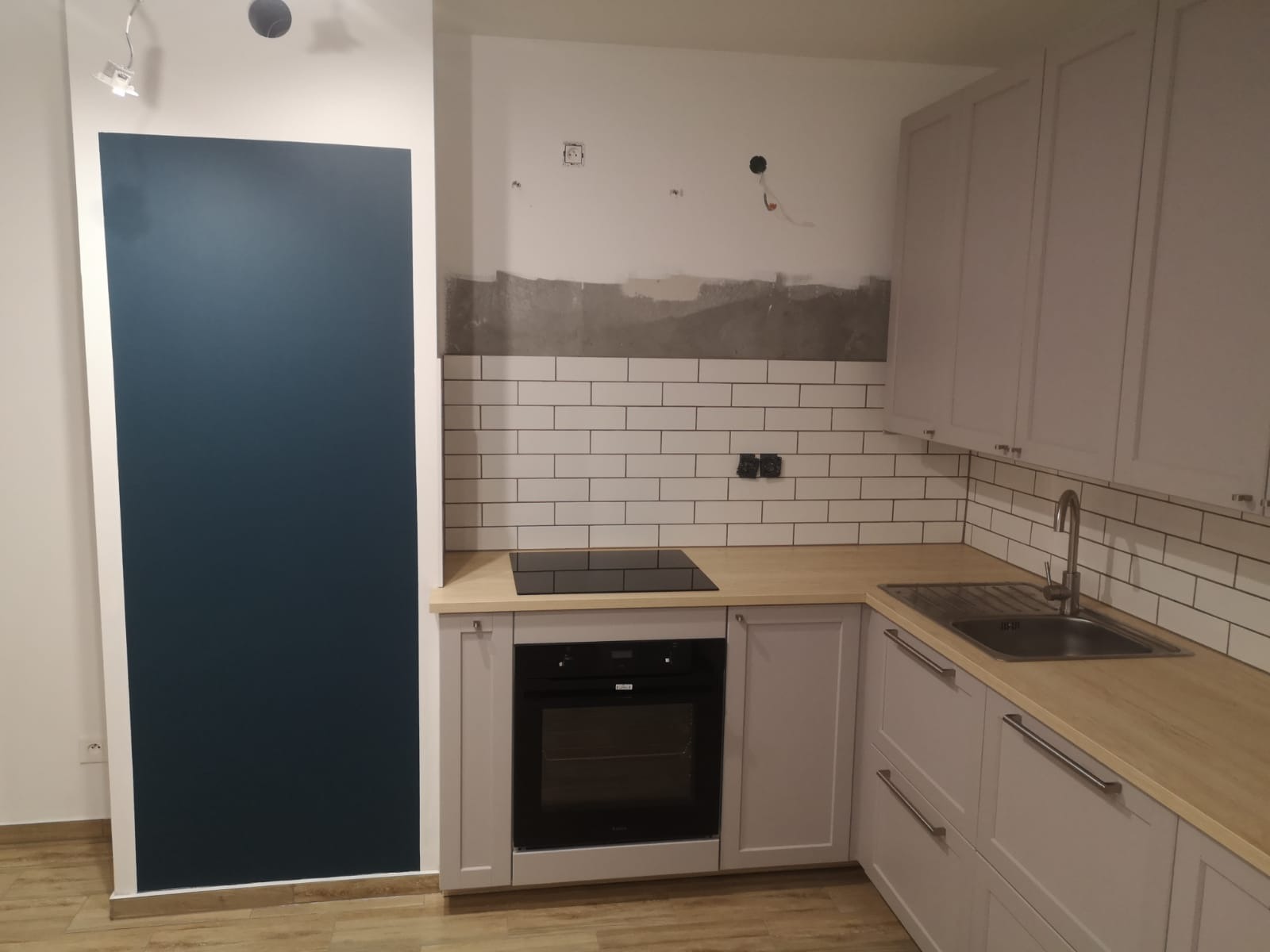Fragment kuchni w trakcie wykańczania: białe płytki metra na ścianie, szare szafki kuchenne z drewnianym blatem, niebieska ściana obok, widoczne ślady prac na ścianie nad płytkami.