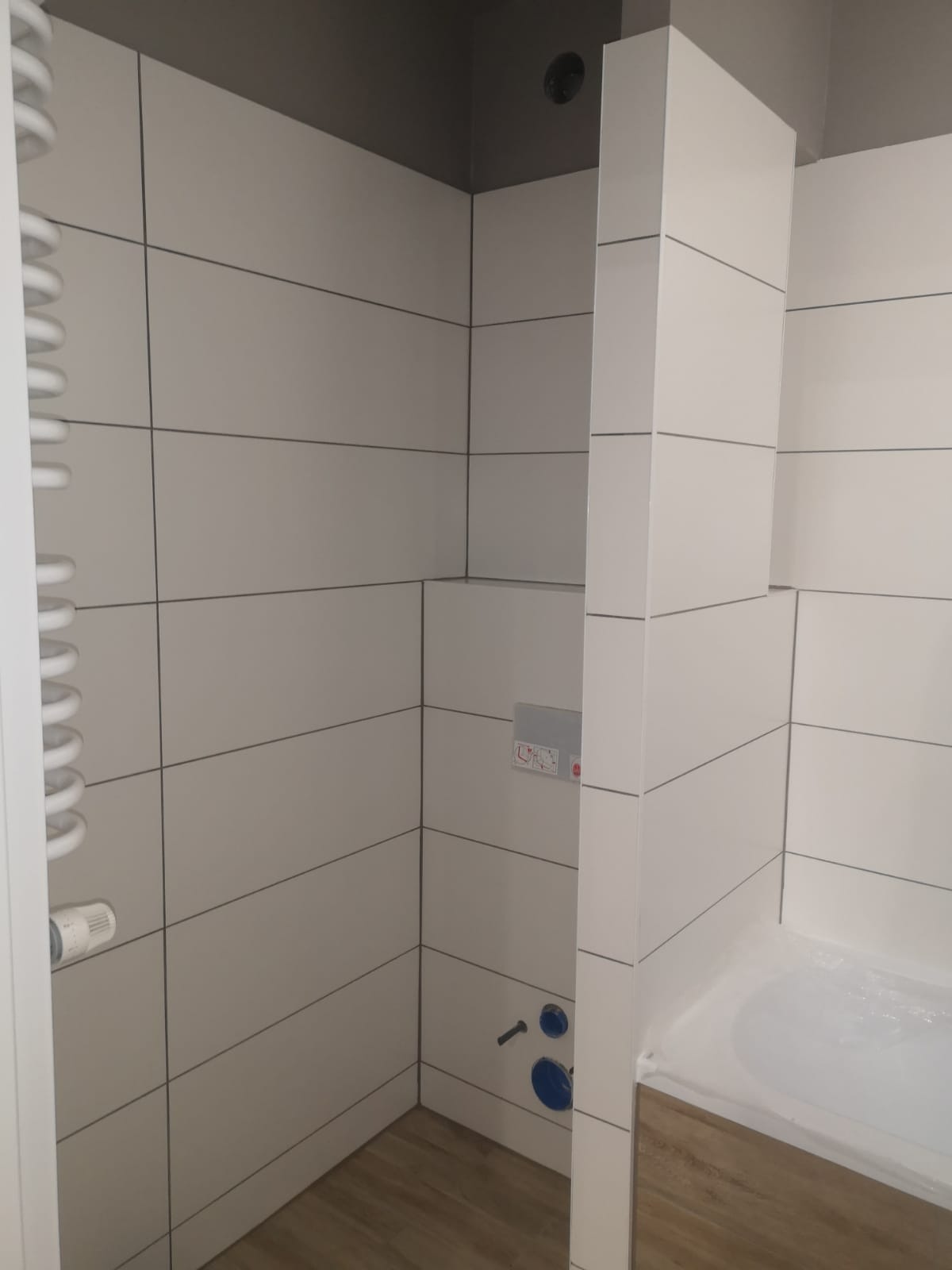 Wykończona łazienka z białymi, poziomymi płytkami, grzejnikiem drabinkowym po lewej stronie i drewnopodobną podłogą, widoczny fragment stelaża podtynkowego WC.