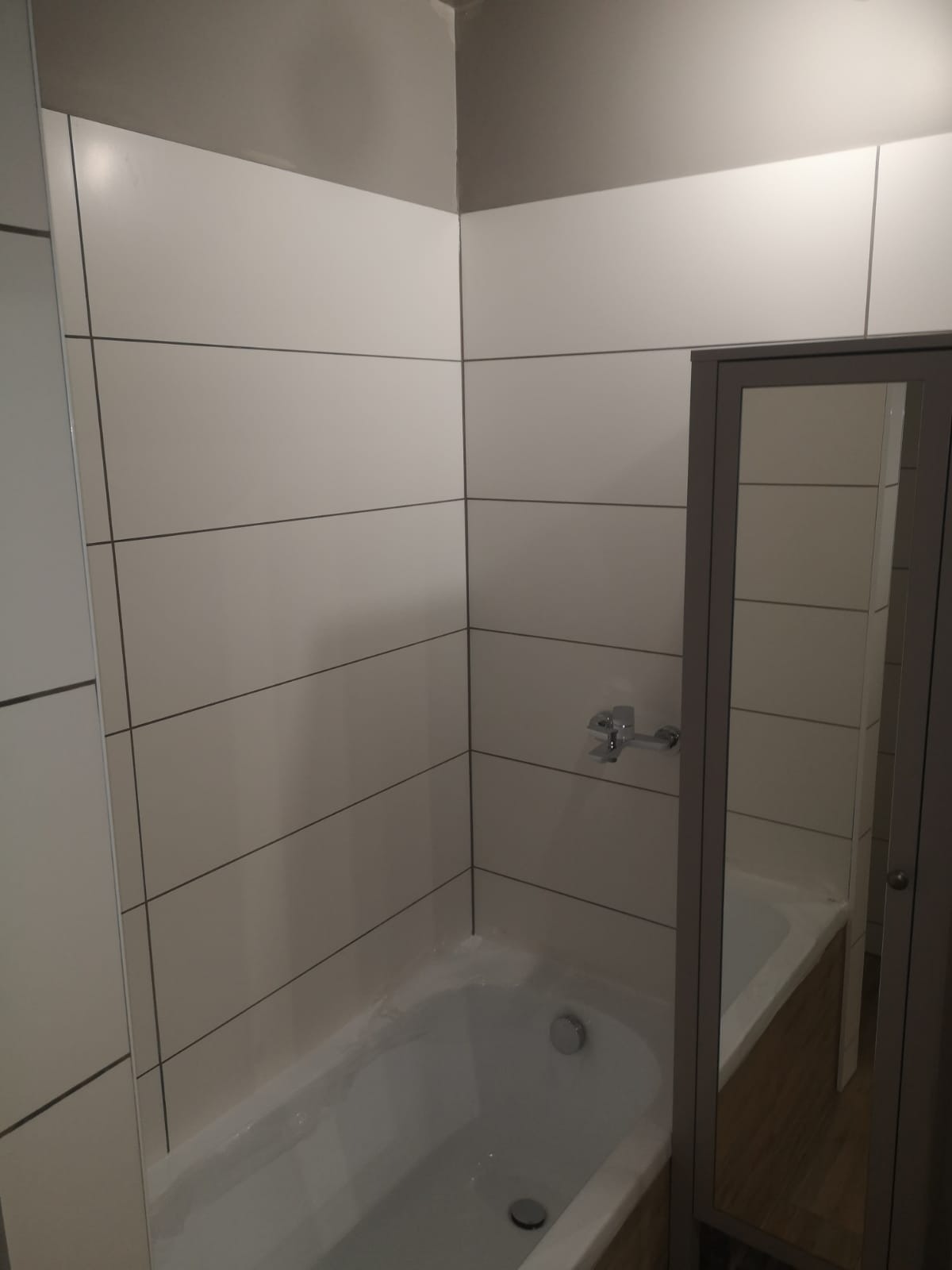 Wykończona łazienka z białymi, poziomymi płytkami i ciemnoszarą fugą, widoczna bateria wannowa i częściowo szafka z lustrem.