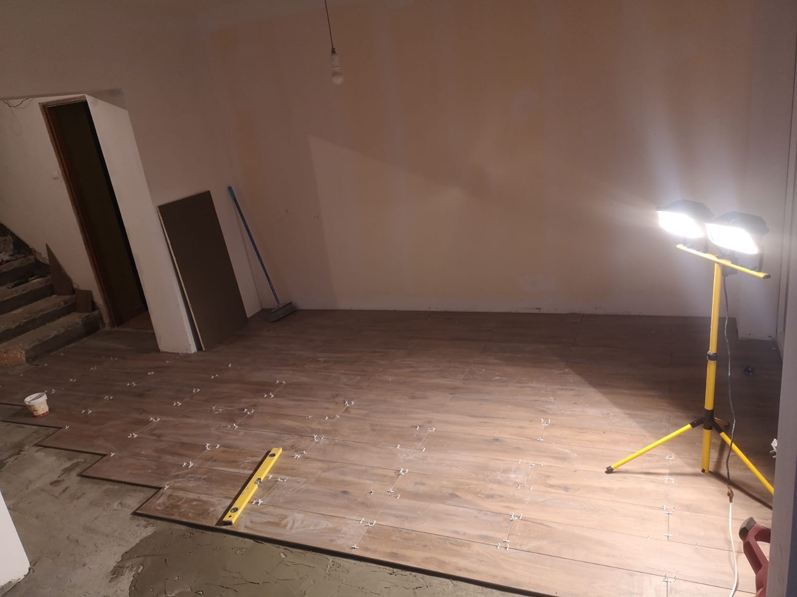Proces układania brązowych płytek podłogowych imitujących drewno, z użyciem krzyżyków dystansowych i poziomicy, oświetlony przenośną lampą budowlaną na trójnogu, widoczne schody i surowy stan ścian.