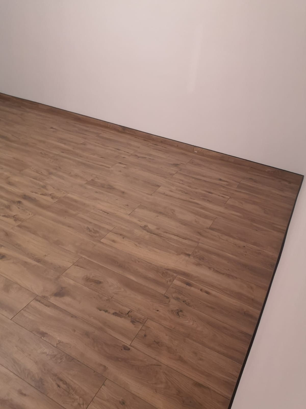 Ułożona brązowa podłoga z paneli imitujących drewno, widoczna czarna listwa przypodłogowa przy białej ścianie.