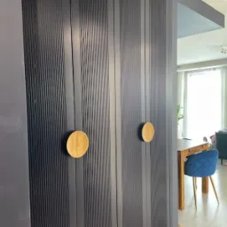 Szara szafa wnękowa z ryflowanymi drzwiami i okrągłymi drewnianymi uchwytami, widok na wnętrze mieszkania z drewnianym stołem i krzesłem.