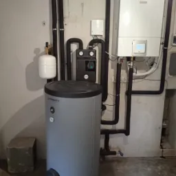 Szary zbiornik buforowy Eternity obok białego pieca grzewczego z wyświetlaczem, czarne rury izolacyjne, betonowa ściana w tle.