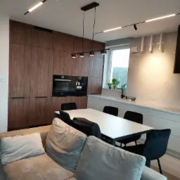 Nowoczesna kuchnia z zabudową z ciemnego drewna, białymi szafkami i blatem, czarnymi krzesłami oraz oświetleniem punktowym i wiszącym, widziana z perspektywy salonu z beżową sofą.