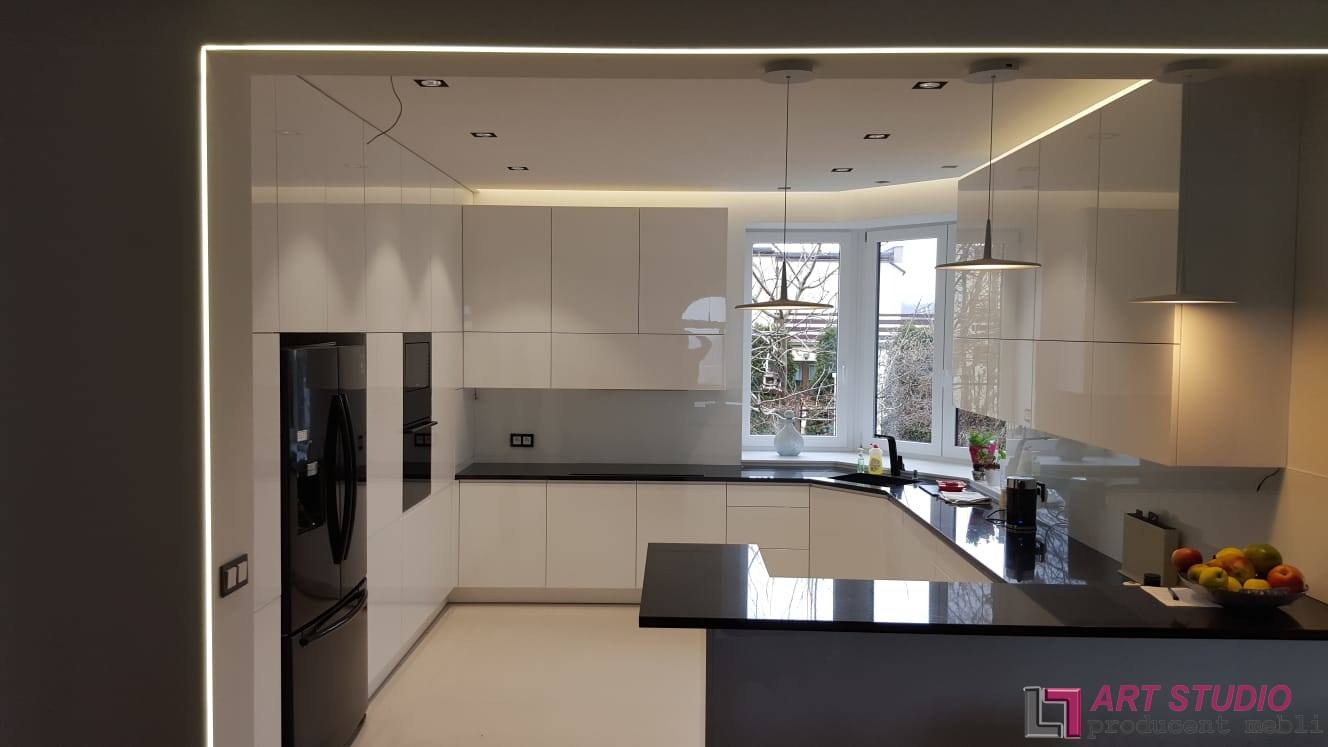 Nowoczesna kuchnia z białymi, lakierowanymi frontami, czarnym blatem i oświetleniem LED podszafkowym, widok na okno z roślinami, minimalistyczne lampy wiszące.