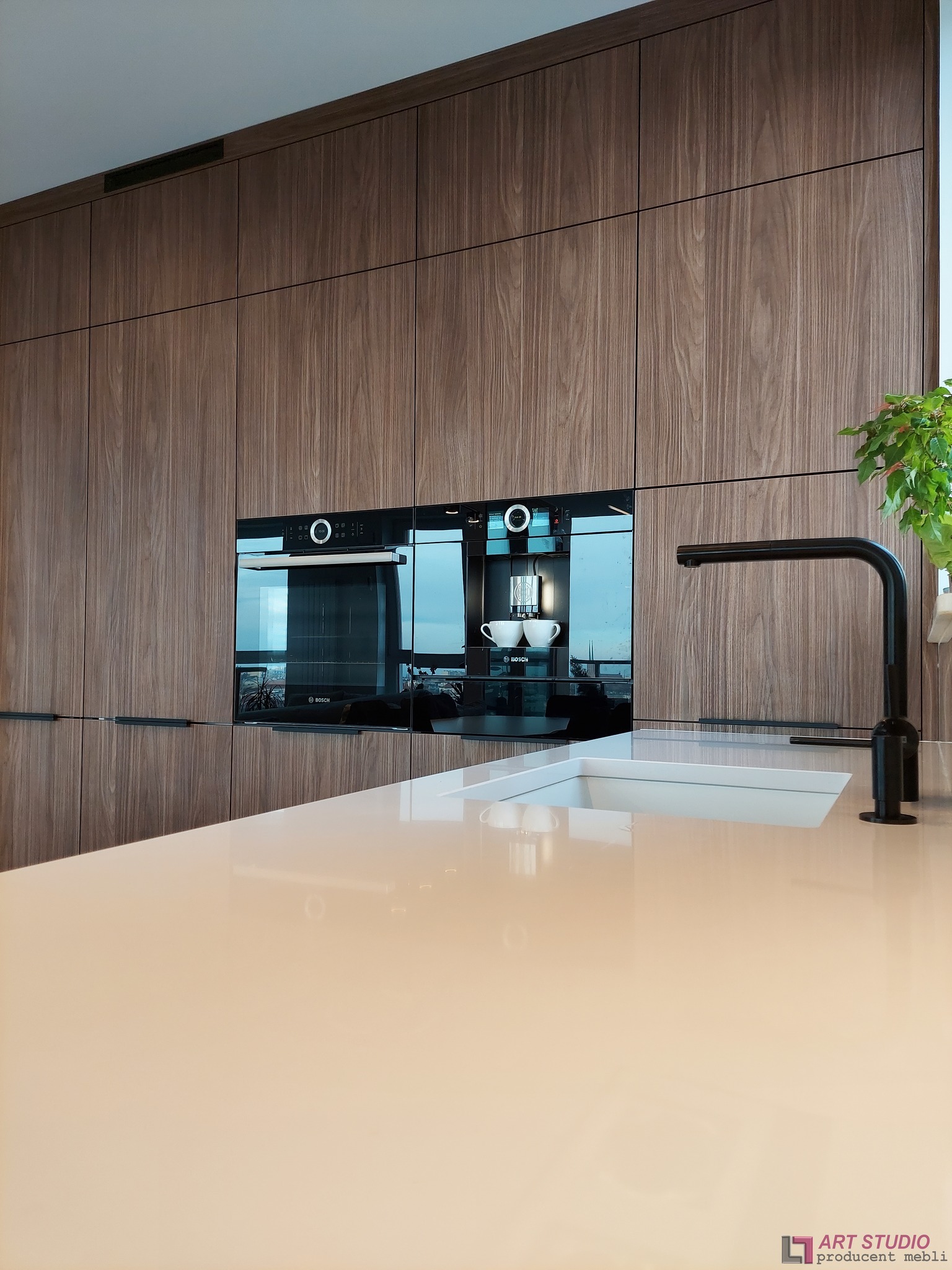 Nowoczesna kuchnia: zabudowa z forniru orzechowego z wbudowanym ekspresem do kawy Bosch, biały blat z wpuszczonym zlewem i czarną baterią.