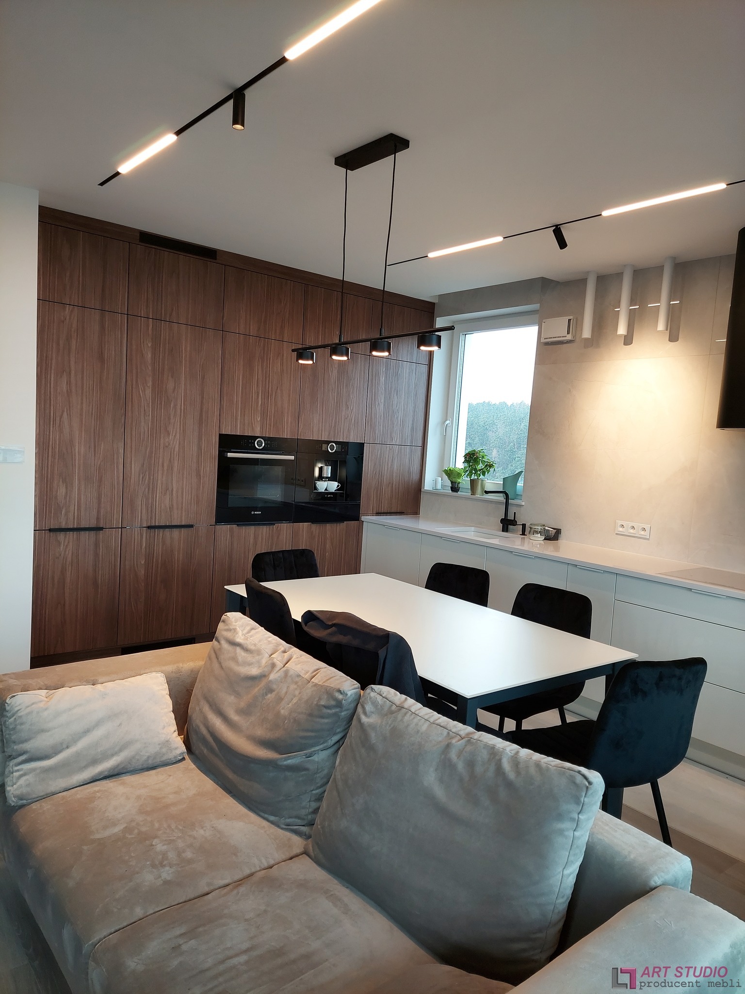 Nowoczesna kuchnia z zabudową z ciemnego drewna, białymi szafkami i blatem, czarnymi krzesłami oraz oświetleniem punktowym i wiszącym, widziana z perspektywy salonu z beżową sofą.