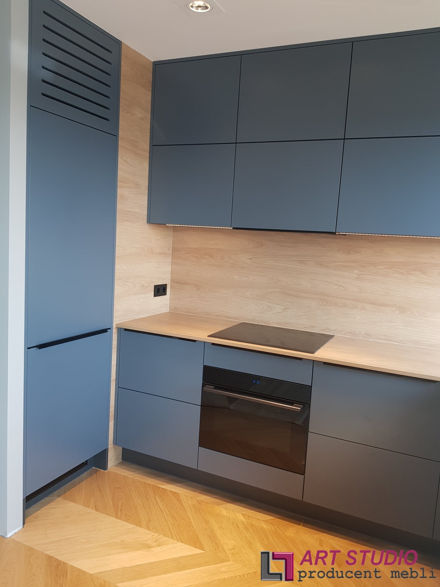 Nowoczesna kuchnia z szarymi, matowymi frontami szafek, drewnianym blatem i podłogą w jodełkę. Widoczny piekarnik z wyświetlaczem i płyta grzewcza. Szafka z żaluzjowymi drzwiczkami po lewej stronie.