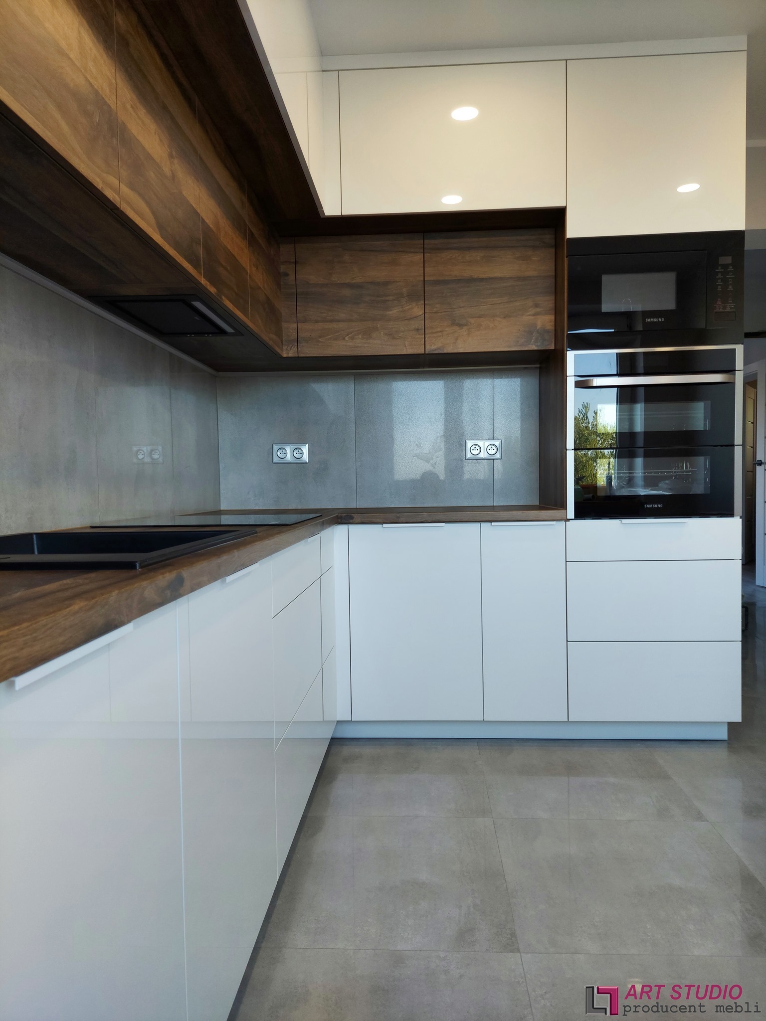 Nowoczesna kuchnia z białymi frontami szafek i blatem z ciemnego drewna, zabudowa z piekarnikiem i mikrofalówką marki Samsung, szara ściana nad blatem i podłoga z płytek w odcieniach szarości.