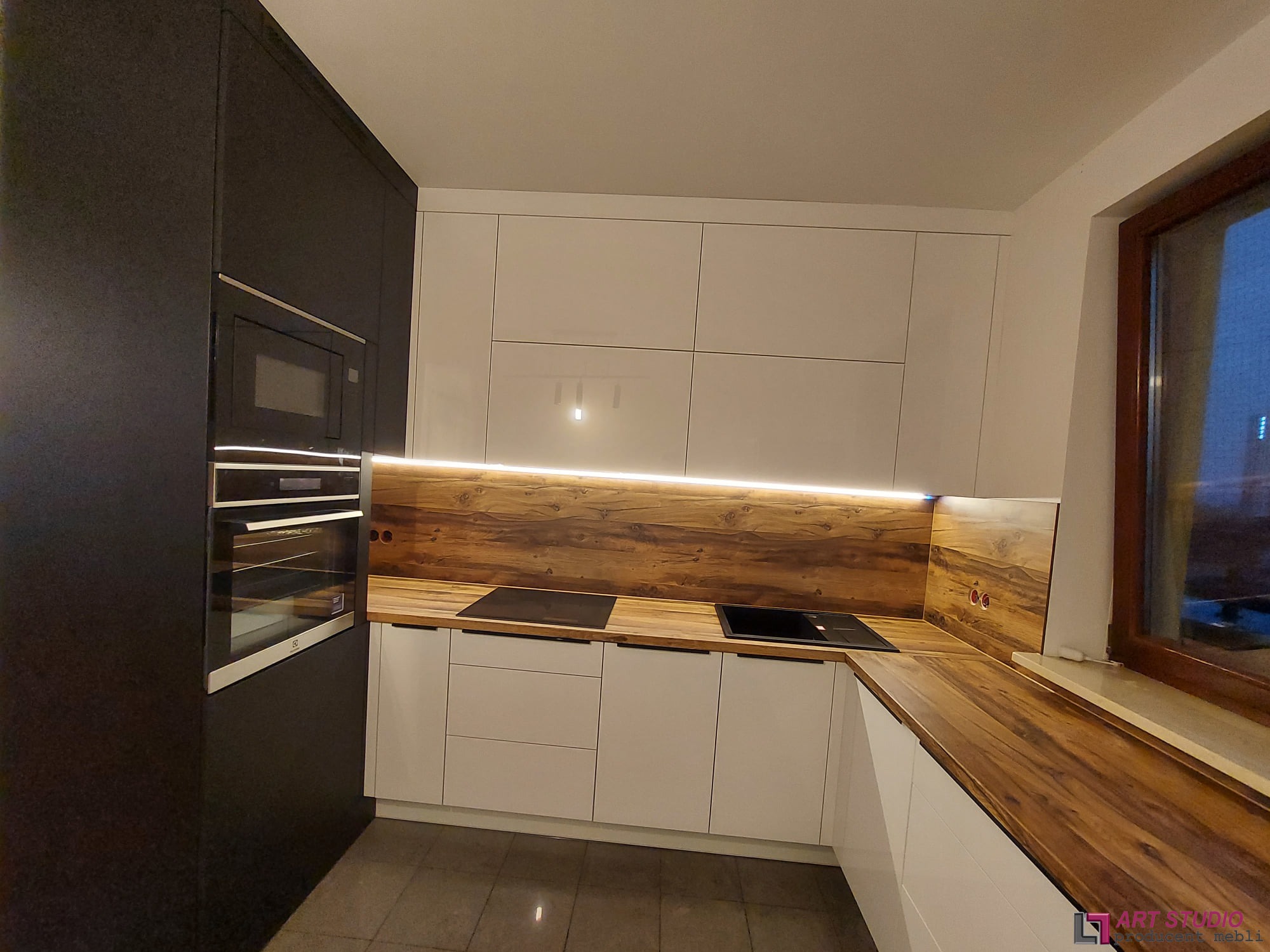 Nowoczesna kuchnia z białymi szafkami i drewnianym blatem, oświetlona listwą LED pod szafkami wiszącymi, z czarnym piekarnikiem w zabudowie i płytą indukcyjną na blacie.