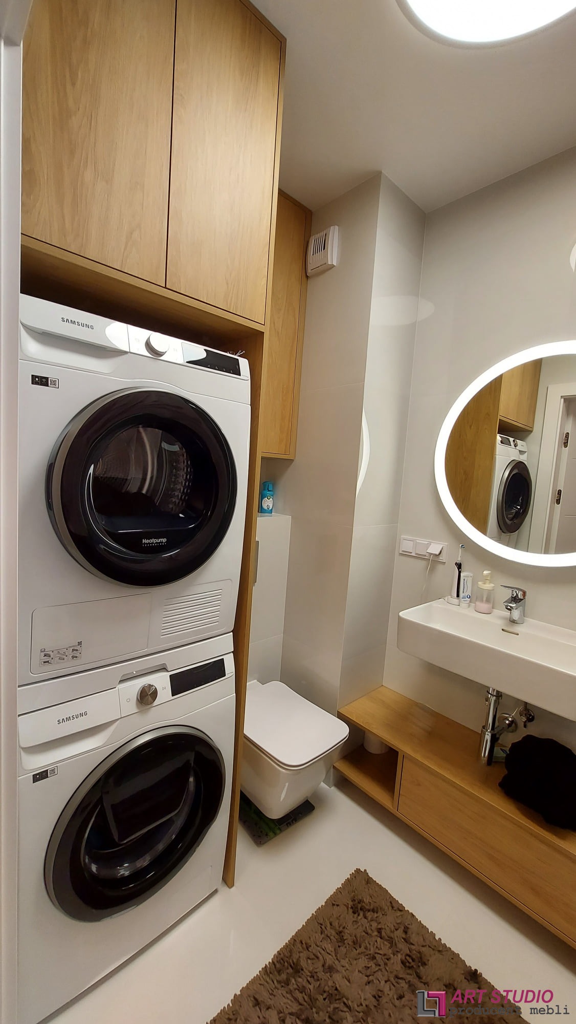 Nowoczesna łazienka z drewnianymi szafkami, pralką i suszarką Samsung w kolumnie, owalnym lustrem z podświetleniem LED oraz brązowym dywanikiem.