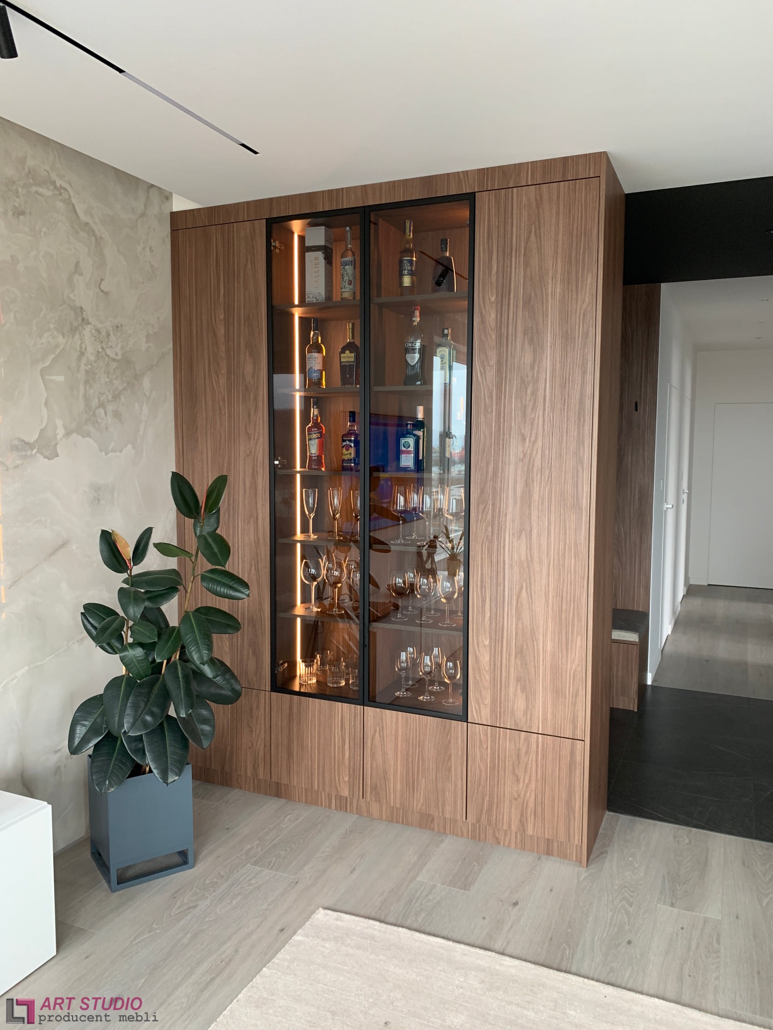 Nowoczesna zabudowa salonu z drewnianą szafą witrynową z podświetleniem LED, eksponującą kolekcję alkoholi i kieliszków, obok marmurowej ściany i rośliny doniczkowej w szarym kwietniku.