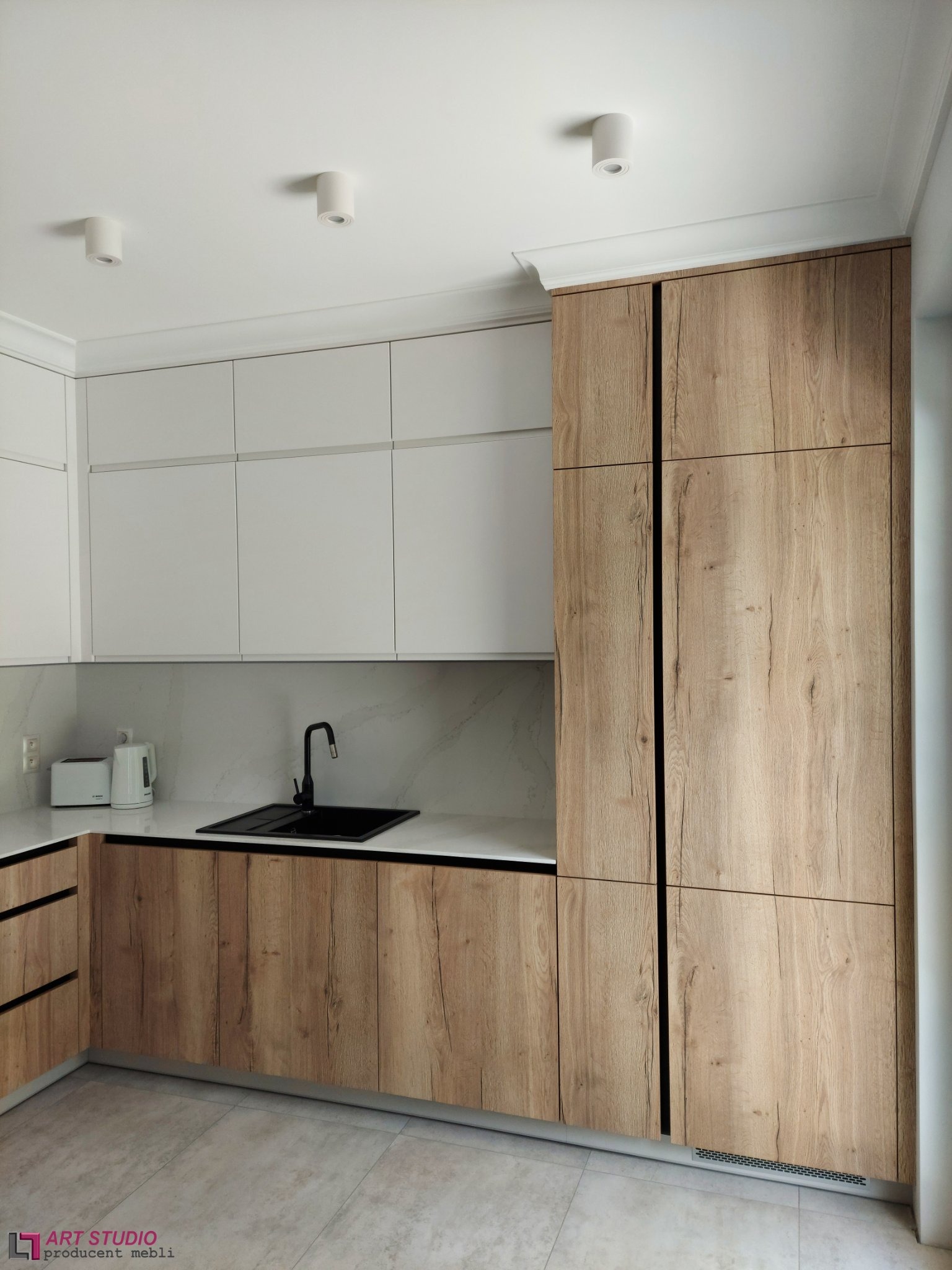 Nowoczesna kuchnia z drewnianymi frontami szafek dolnych i wysokiej zabudowy, oraz białymi szafkami górnymi, czarny zlew i bateria.