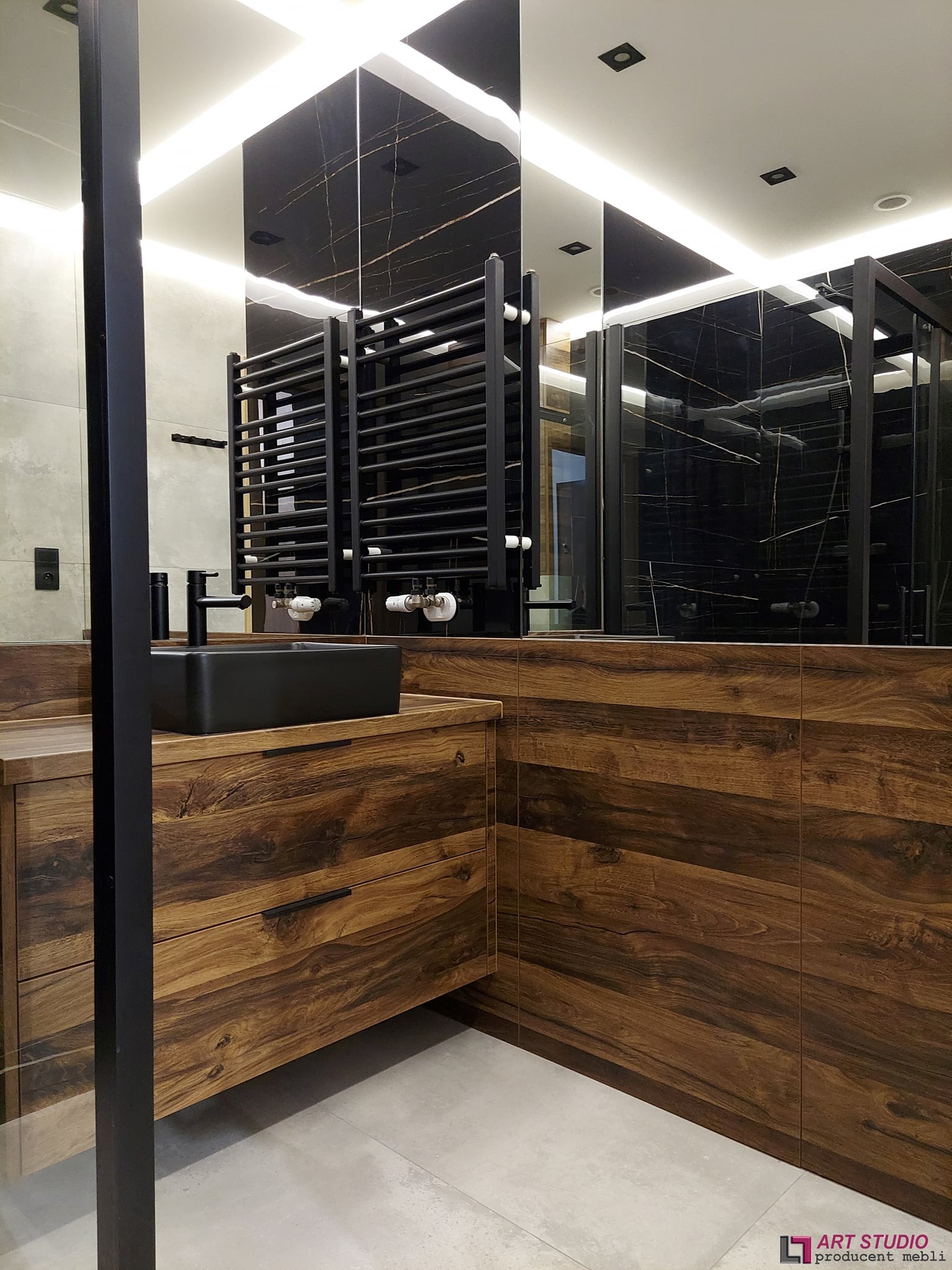 Nowoczesna łazienka z drewnianą szafką pod umywalkę, czarną armaturą i grzejnikiem drabinkowym na tle czarnej ściany z marmurowym wzorem odbijającej się w lustrze.