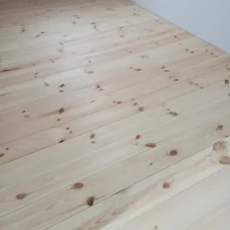 Jasne, świeżo ułożone deski podłogowe z widocznymi sękami, ułożone równolegle do ściany, w rogu pokoju.
