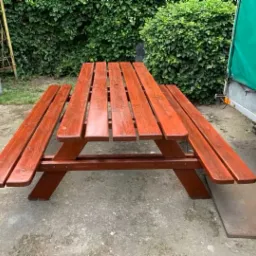 Drewniany stół piknikowy z ławkami, pomalowany na czerwono-brązowy kolor, ustawiony na betonowym podłożu w ogrodzie, z widoczną przyczepką samochodową w tle.