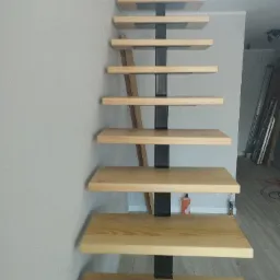 Nowoczesne schody z drewnianymi stopniami i czarną metalową konstrukcją nośną, widok od dołu, w tle widoczne materiały budowlane.