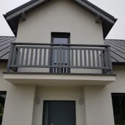 Dwukondygnacyjny dom z balkonem z szarą balustradą i szarą stolarką okienną oraz drzwiami wejściowymi w tym samym odcieniu szarości.