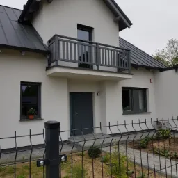 Nowoczesny dom jednorodzinny z szarym dachem, balkonem z drewnianą balustradą i ogrodzeniem panelowym, w trakcie zagospodarowywania terenu.