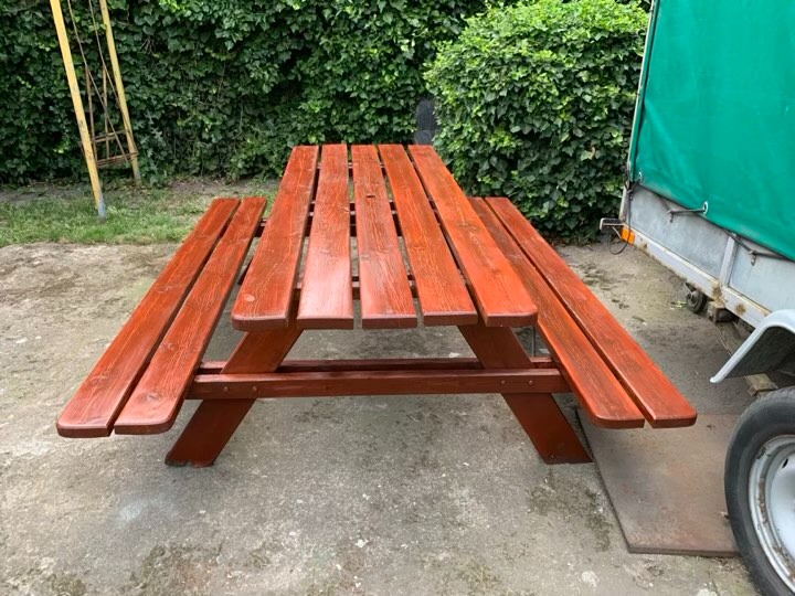 Drewniany stół piknikowy z ławkami, pomalowany na czerwono-brązowy kolor, ustawiony na betonowym podłożu w ogrodzie, z widoczną przyczepką samochodową w tle.