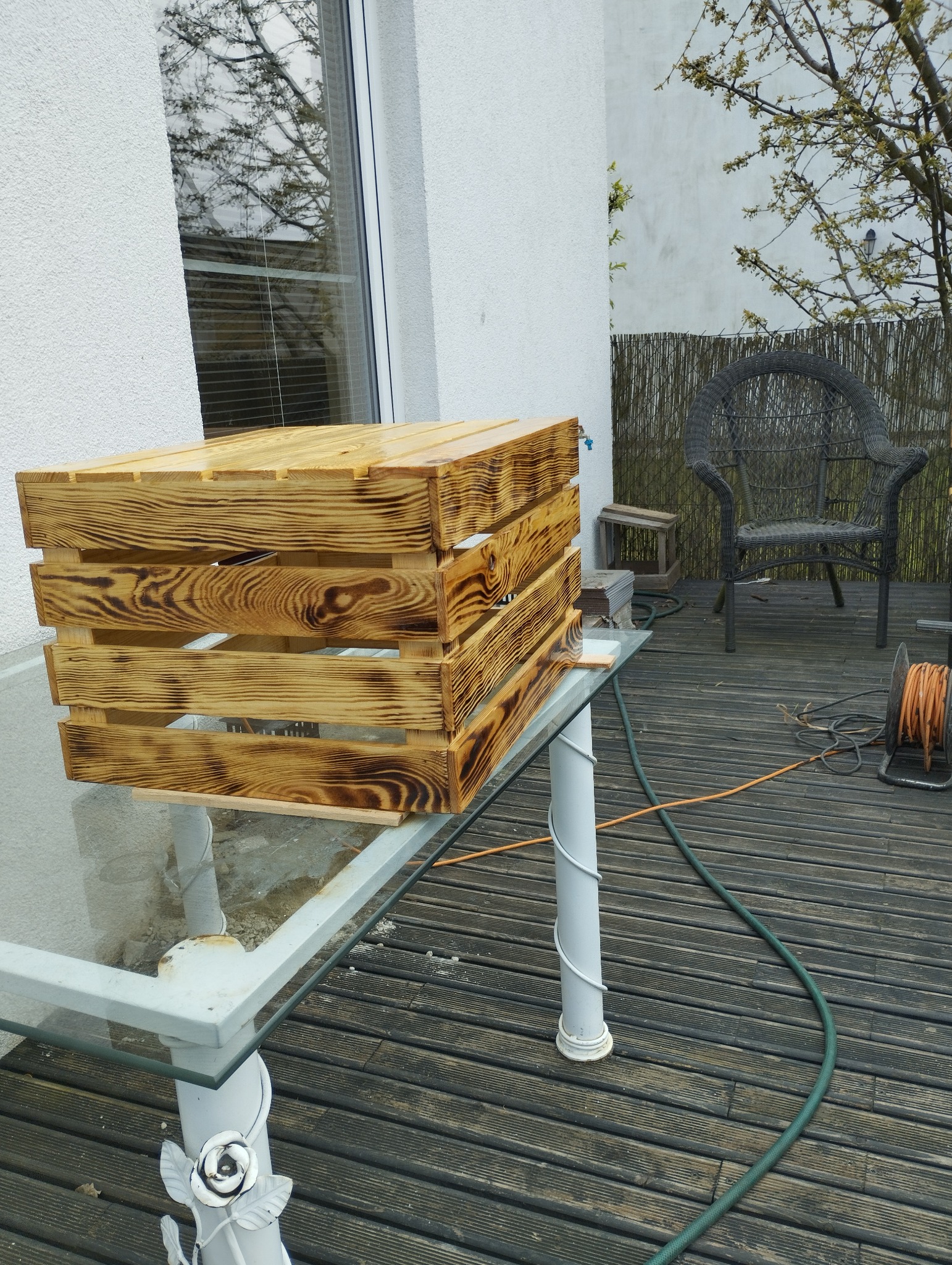 Drewniana skrzynka z efektem opalania, stojąca na szklanym stole na tarasie z drewnianą podłogą, w tle krzesło wiklinowe i fragment ogrodu.