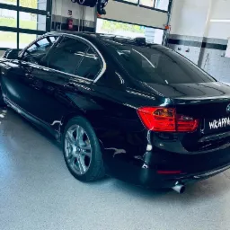 Błyszczący, czarny sedan BMW 320i z przyciemnionymi szybami, zaparkowany w dobrze oświetlonym garażu z podnoszoną bramą i szarą podłogą.