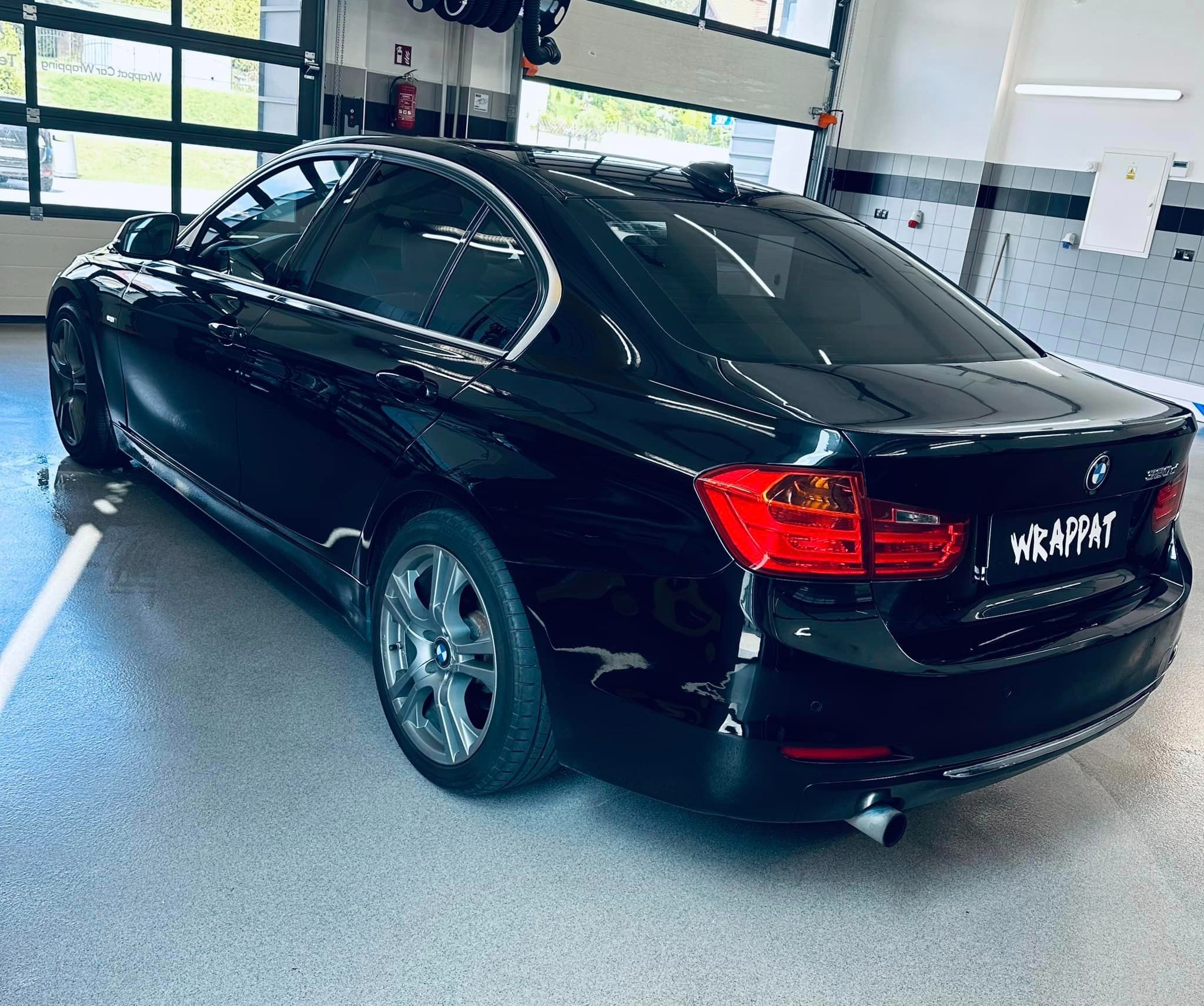 Błyszczący, czarny sedan BMW 320i z przyciemnionymi szybami, zaparkowany w dobrze oświetlonym garażu z podnoszoną bramą i szarą podłogą.