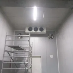 Wnętrze chłodni w Sochaczewie z zamontowanym agregatem chłodniczym z trzema wentylatorami oraz wentylatory ścienne. Widoczne rusztowanie.