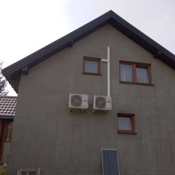 Damian Ziółkowski - Dwa klimatyzatory zamontowane na szarej ścianie domu jednorodzinnego z brązowymi oknami, z białą rurą odprowadzającą skropliny i panelem słonecznym opartym o ścianę, na tle zielonego trawnika.