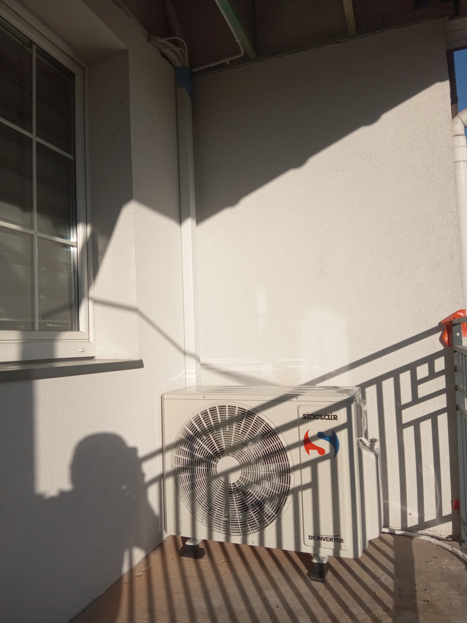 Zewnętrzna jednostka klimatyzacji Sinclair DC Inverter zamontowana na elewacji budynku, widoczna instalacja elektryczna i rury odprowadzające skropliny, cień barierki i osoby robiącej zdjęcie.