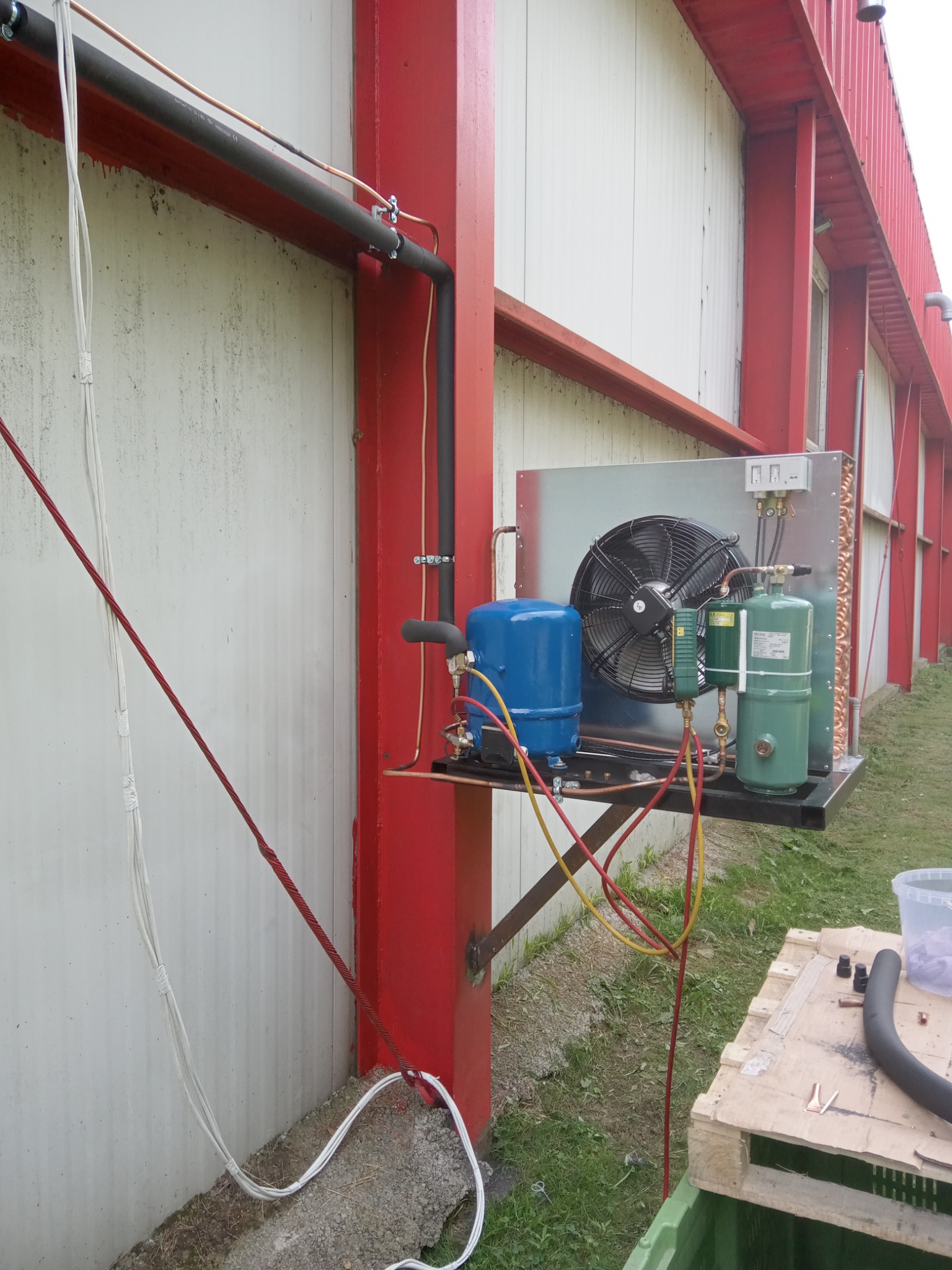 Zewnętrzna jednostka klimatyzacyjna zamontowana na czerwonym słupie konstrukcyjnym budynku, widoczne przewody, wentylator, butle i elementy instalacji chłodniczej.