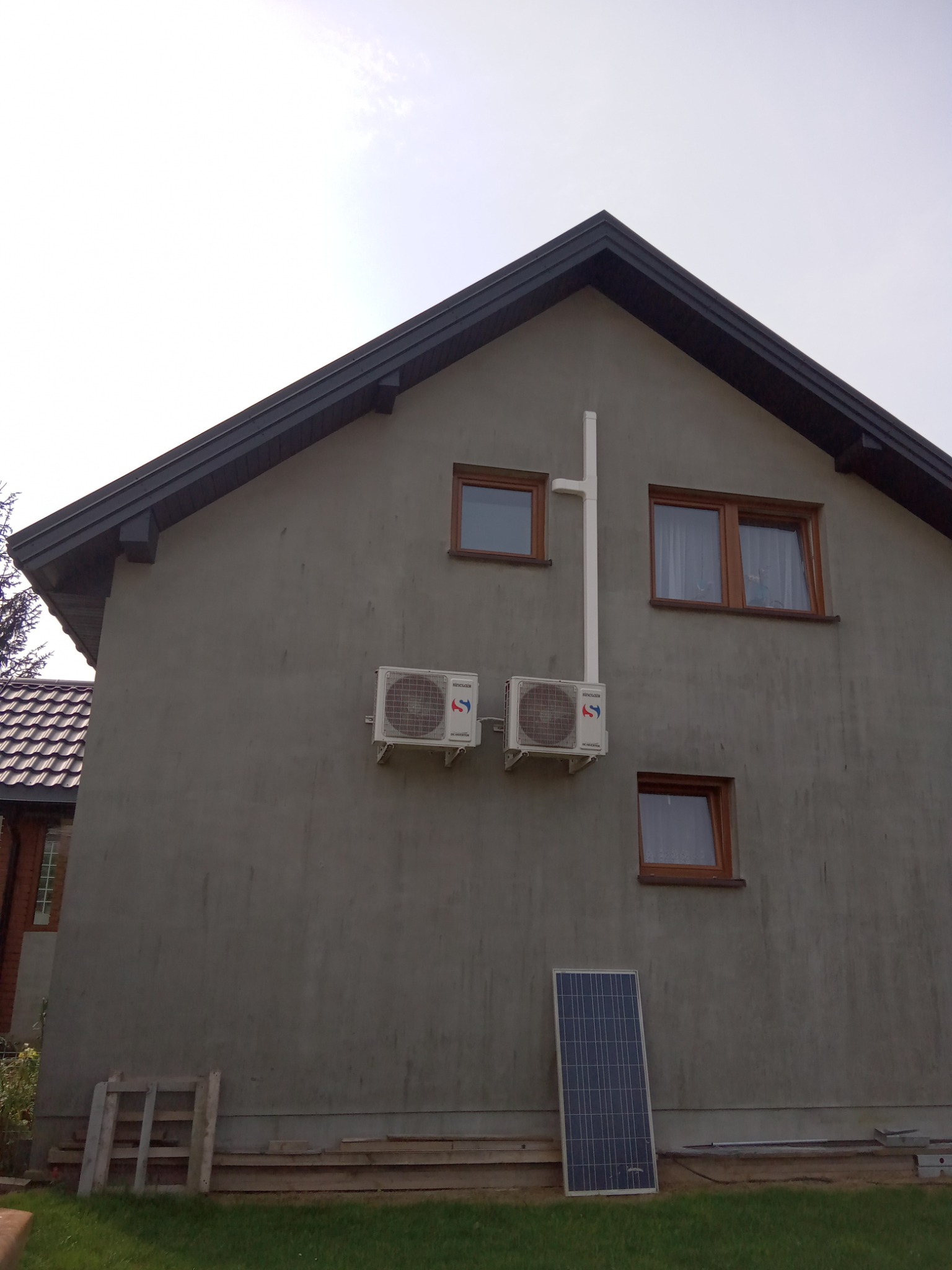 Dwa klimatyzatory zamontowane na szarej ścianie domu jednorodzinnego z brązowymi oknami, z białą rurą odprowadzającą skropliny i panelem słonecznym opartym o ścianę, na tle zielonego trawnika.
