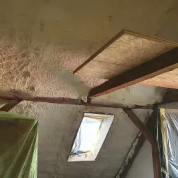 Poddasze z widoczną izolacją pianką natryskową na skośnych ścianach i suficie, drewniane belki konstrukcyjne oraz okno dachowe zabezpieczone folią malarską.