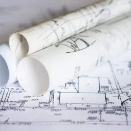 Dwa zwinięte plany architektoniczne leżące na rozłożonym planie z odręcznymi notatkami i wymiarami, widoczne numery działek i nazwy ulic.