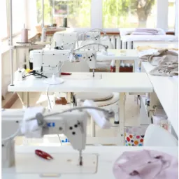 Jasne wnętrze szwalni z kilkoma białymi maszynami do szycia JUKI, widok z perspektywy, na stole leży niedokończone jasnoróżowe ubranko dziecięce.