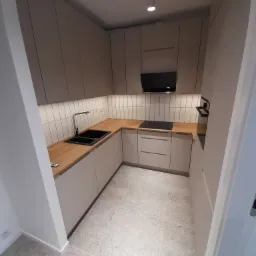 Nowoczesna kuchnia w odcieniach beżu i brązu z drewnianym blatem, czarnym zlewem i płytą indukcyjną, oświetlona punktowymi lampami sufitowymi i pod szafkami.