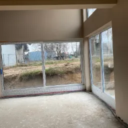 Wnętrze w stanie deweloperskim po nałożeniu gładzi szpachlowej na ścianach, z widocznymi dużymi oknami przesuwnymi i przewodami elektrycznymi przy podłodze.