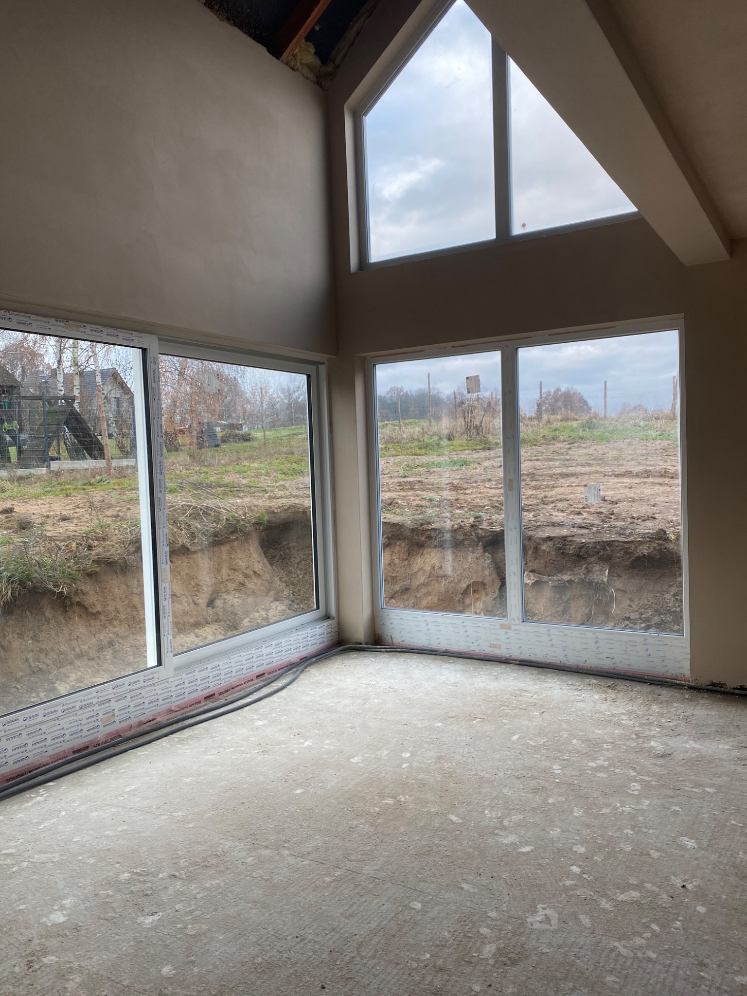 Surowe wnętrze domu po nałożeniu tynków, z dużymi oknami wychodzącymi na krajobraz z wykopem w ziemi. Widoczne zaciągnięte ściany i sufit oraz surowa podłoga.